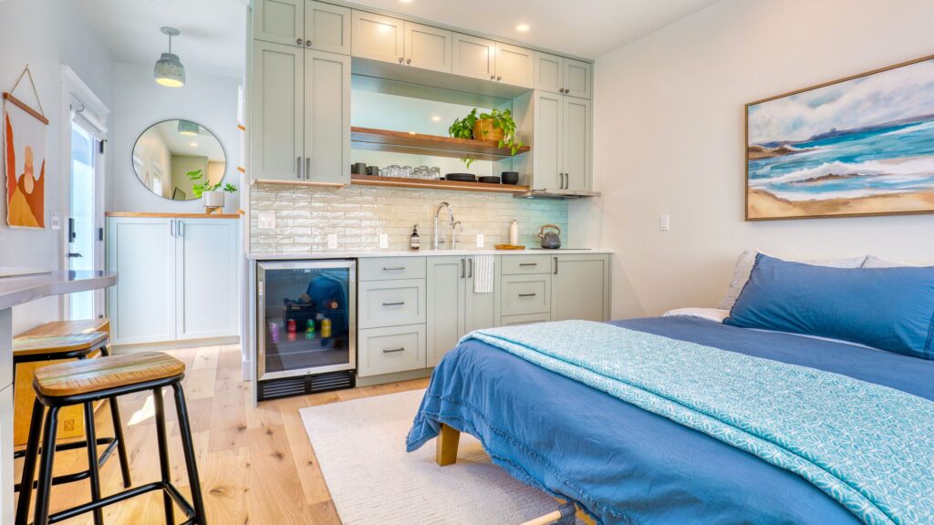 Interior of a DADU in Seattle with sage kitchenette cabinets, open wood shelving, a beverage fridge, and a coastal-themed bedroom area with blue and teal bedding.