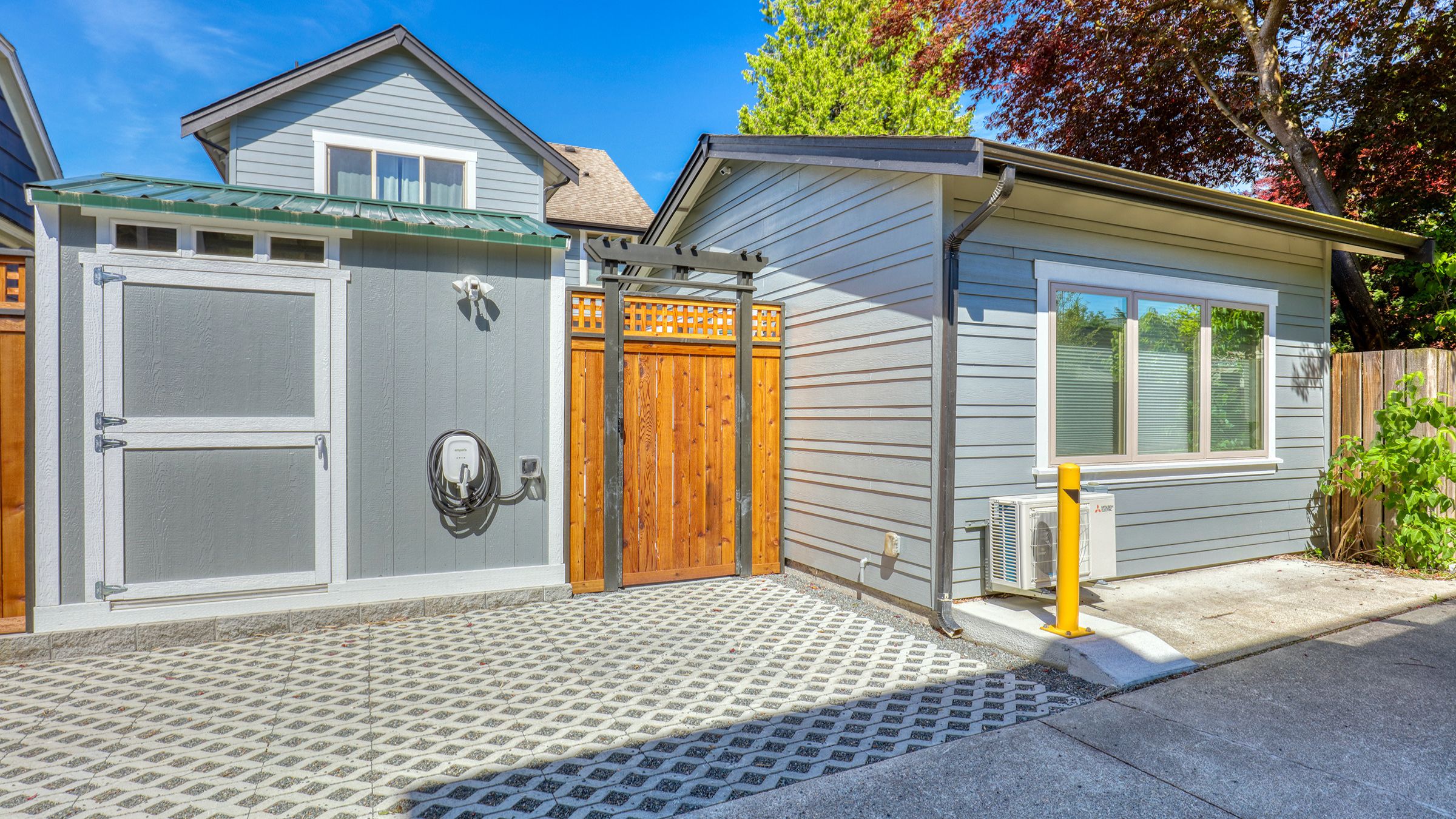 A gray DADU in Seattle with a permeable paver driveway, EV charger, cedar gate, and mini-split heat pump, set beside a storage shed with a green metal roof.